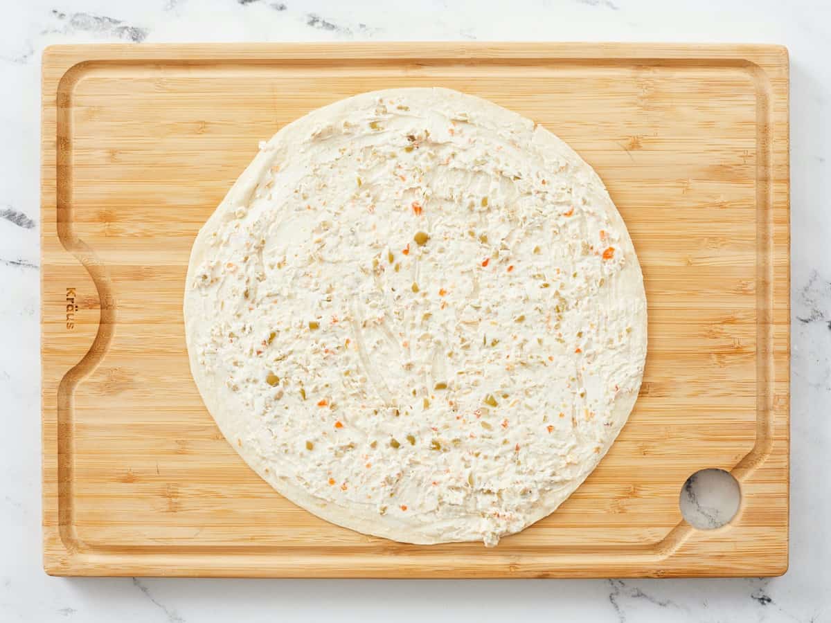 An extra large burrito shell smeared from edge to edge with a savory cream cheese spread, sitting on a wooden cutting board with a white marble background. 
