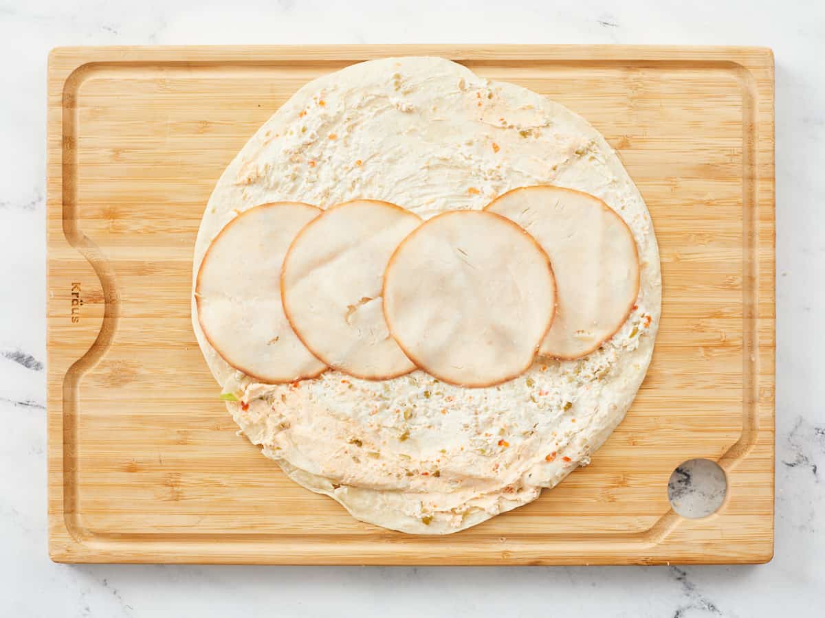 A large tortilla covered with savory cream cheese spread, and in the middle of the tortilla there are four turkey slices line up and overlapping slightly. The tortilla is sitting on a wooden cutting board against a background of white marble.